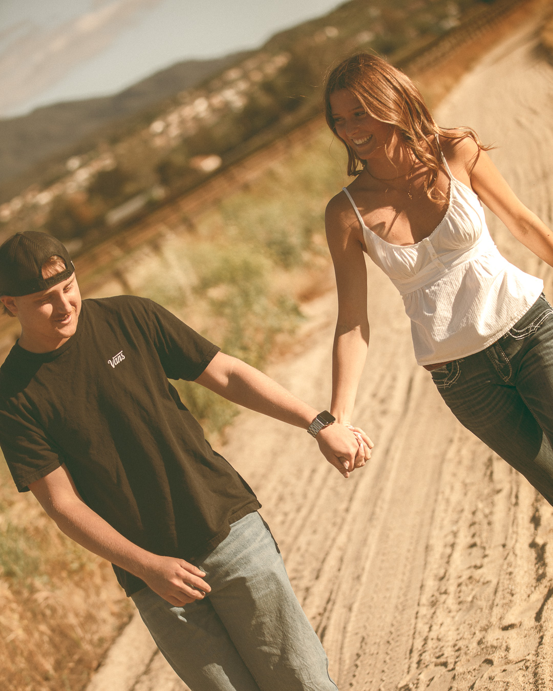 Couple laughing and holding hands as they walk down a sunlit dirt road